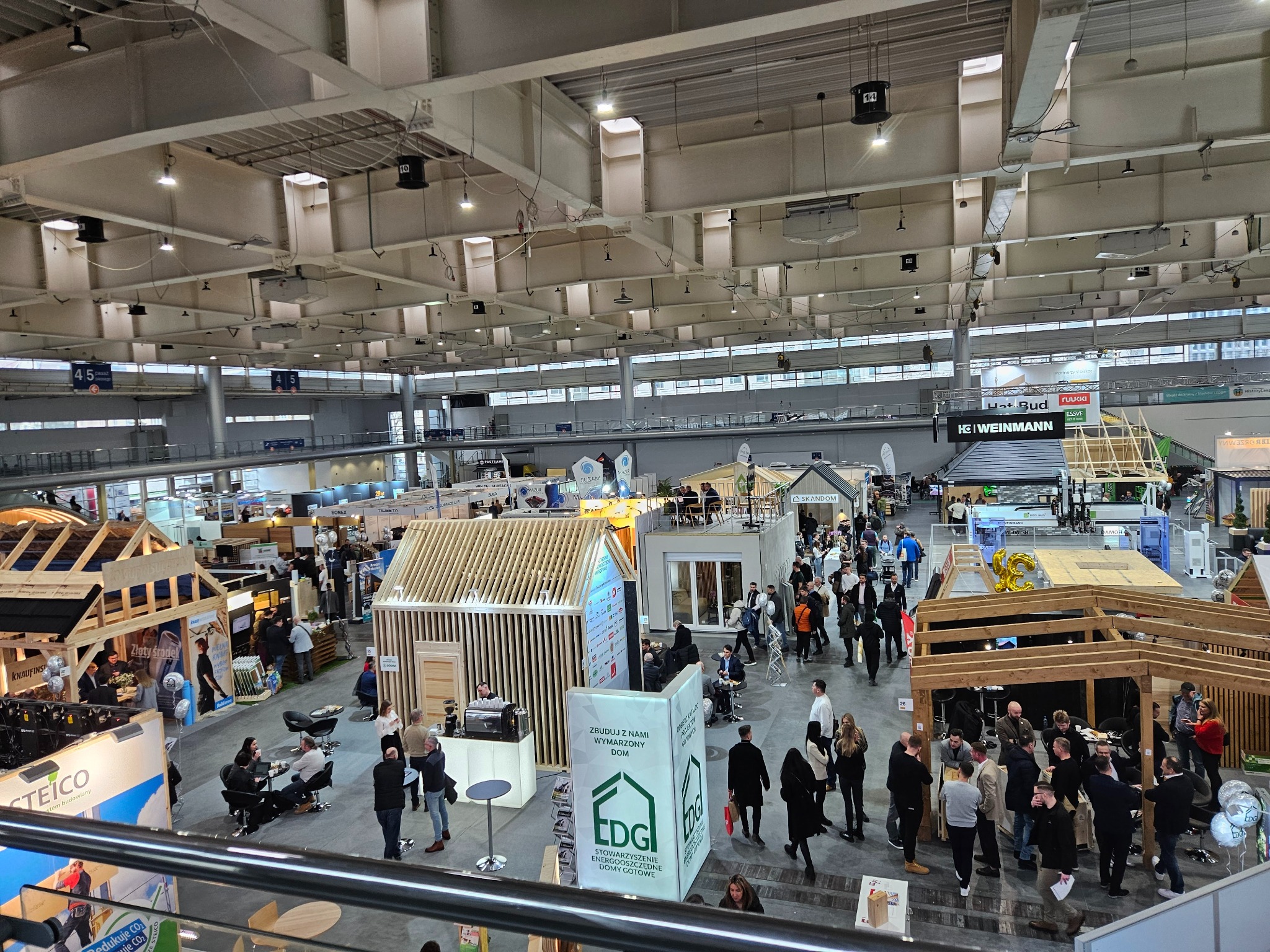 Widok z góry na halę targową z ekspozycjami domów modułowych i szkieletowych, liczne osoby zwiedzają stoiska. Jasne oświetlenie, widoczne konstrukcje dachowe hali.