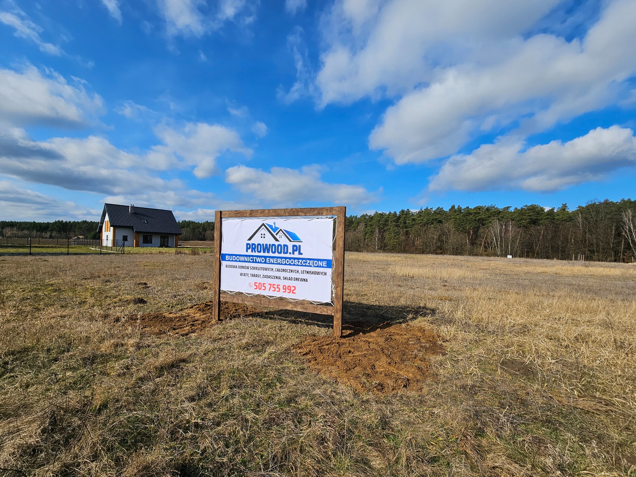Tablica informacyjna Prowood.pl na tle budowanego domu szkieletowego i łąki. Reklama budownictwa energooszczędnego: domy szkieletowe, całoroczne, letniskowe. Numer telefonu.
