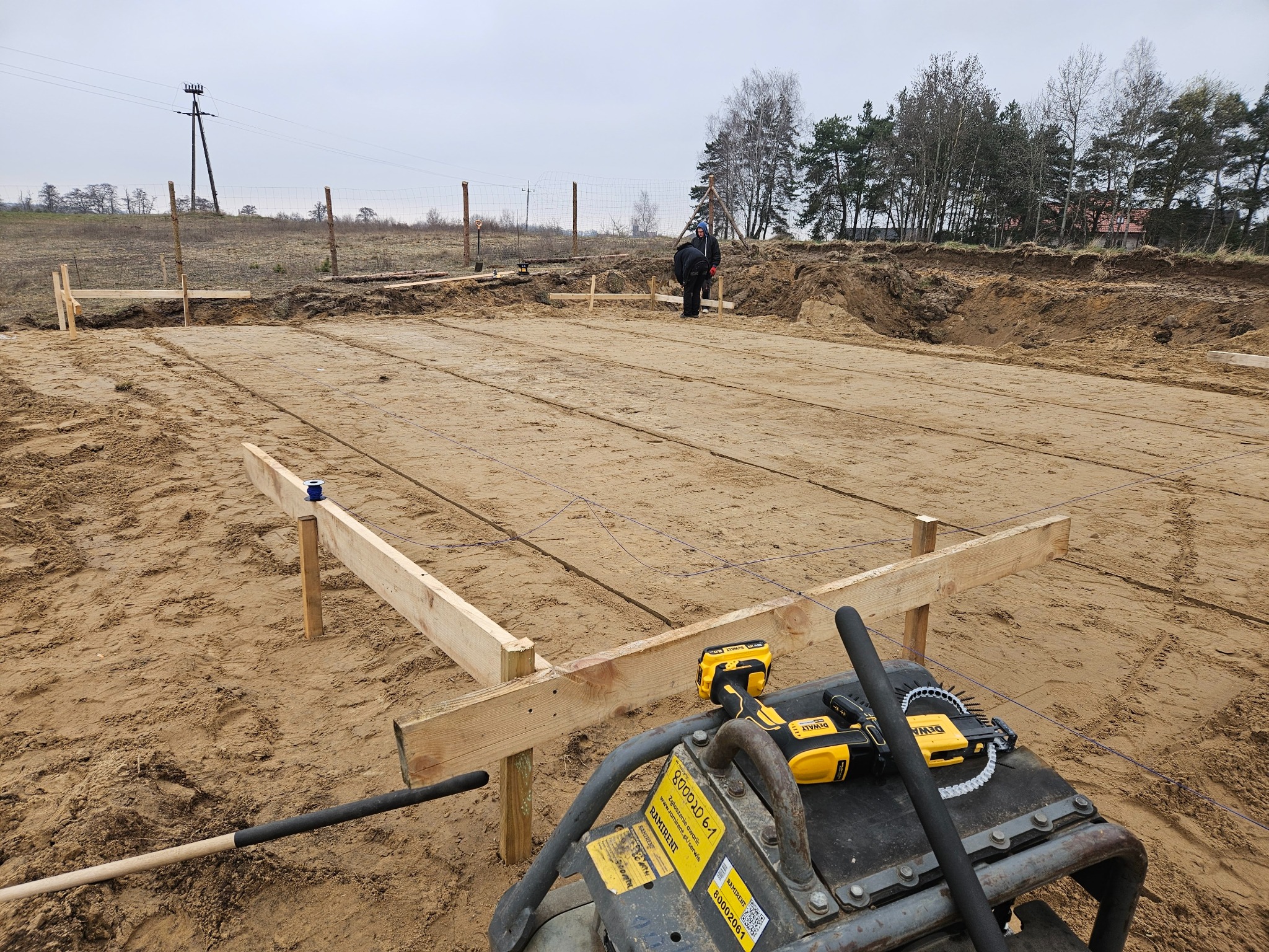Przygotowany teren pod budowę domu: wytyczone fundamenty, drewniane szalunki i sprzęt budowlany na piaszczystym podłożu. Widoczni pracownicy w tle.