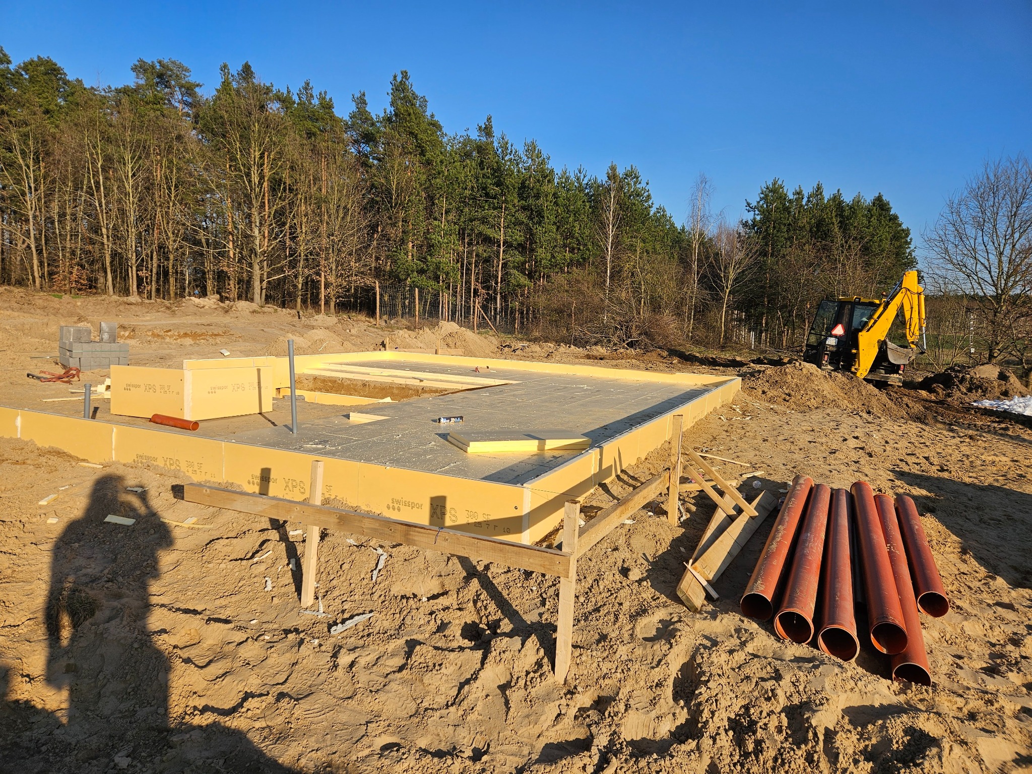 Fundament domu z izolacją XPS, przygotowany pod budowę. Na placu budowy widoczny żółty traktor i rury kanalizacyjne. W tle las i błękitne niebo, cień osoby w lewym rogu.