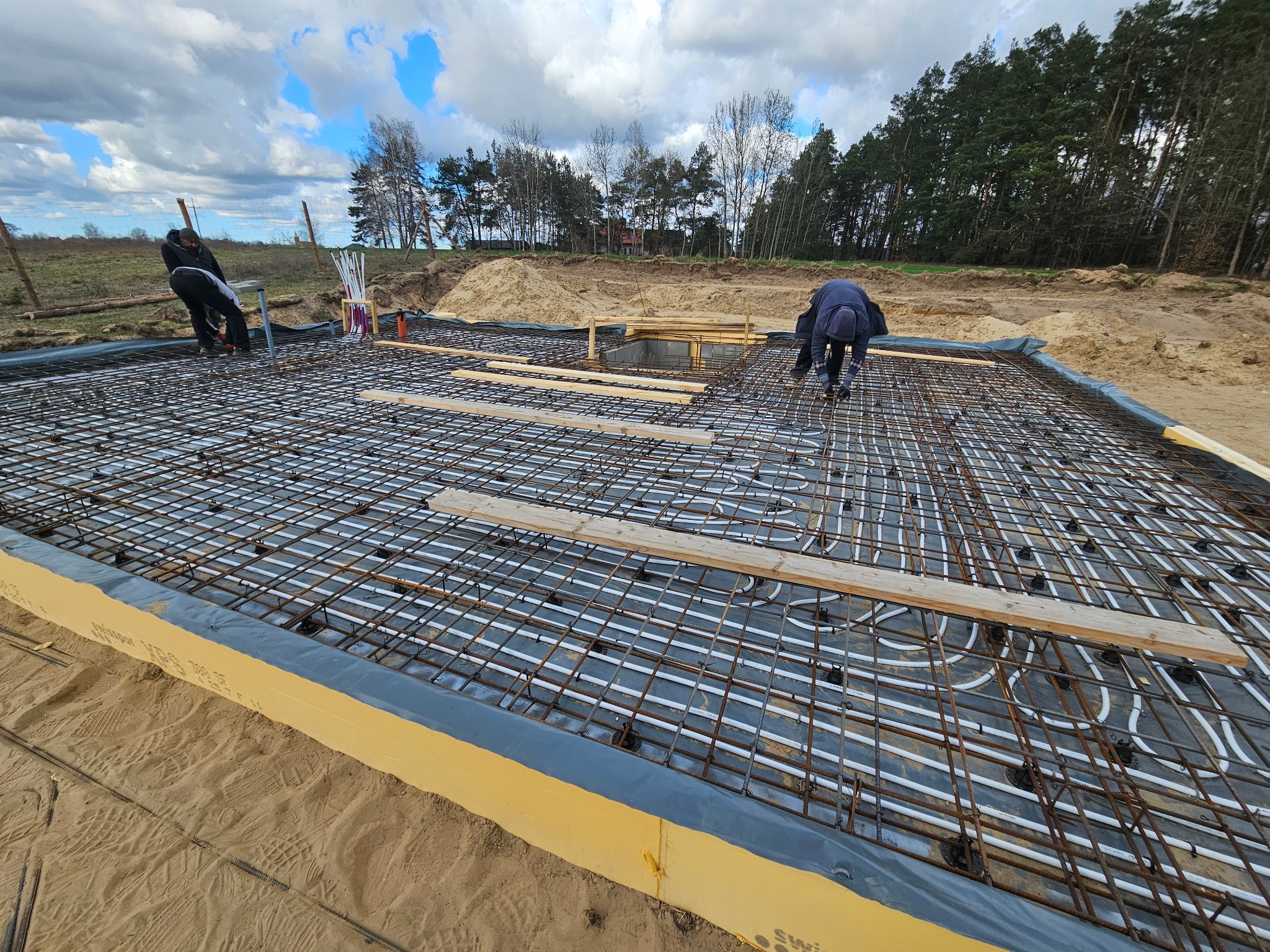 Montaż ogrzewania podłogowego w fundamencie domu szkieletowego. Widoczne rury grzewcze, siatka zbrojeniowa i pracownicy. Fundament zabezpieczony izolacją.