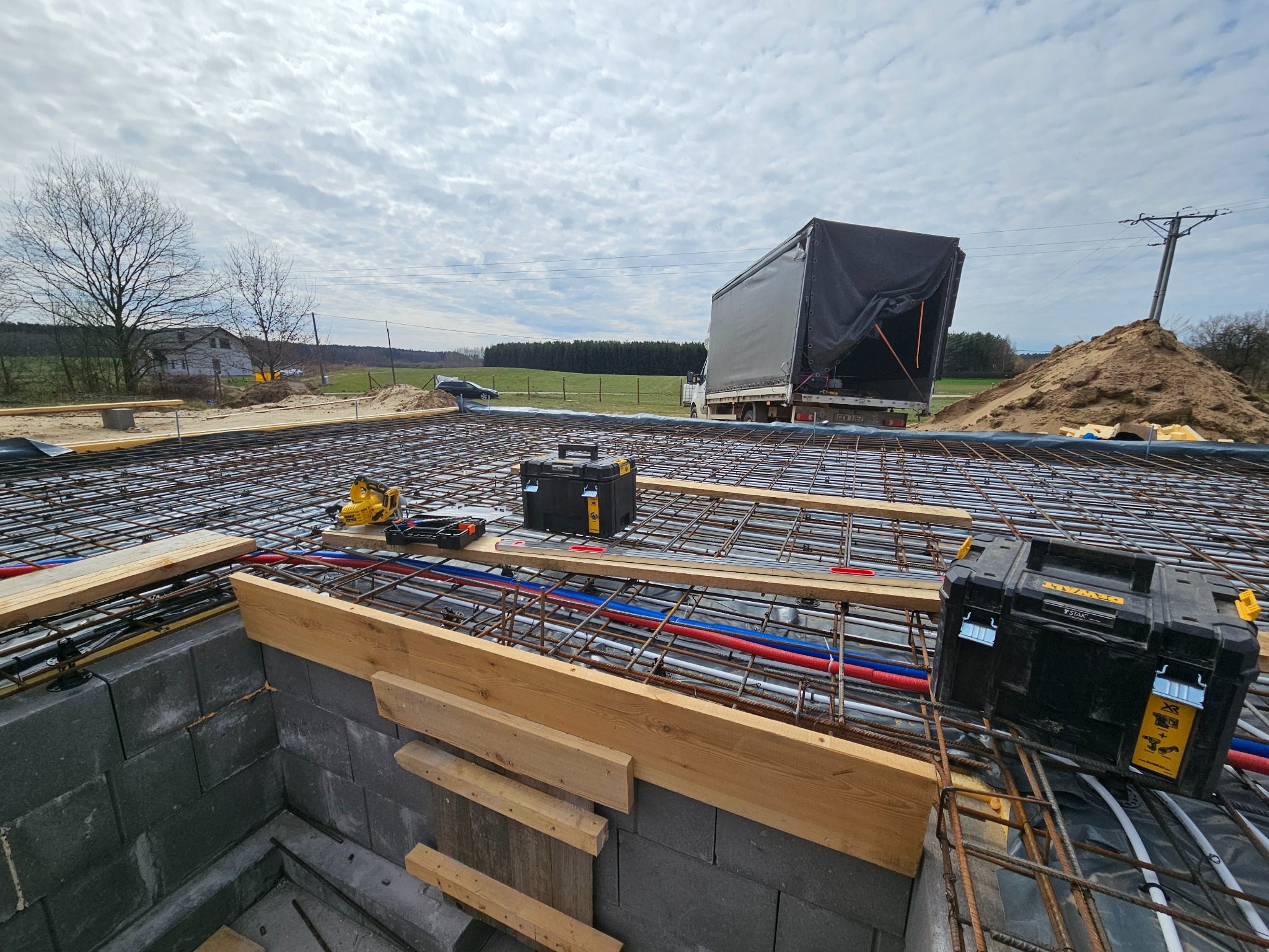 Zbrojenie płyty fundamentowej z instalacją podposadzkową (rury CO i wod-kan) na budowie domu jednorodzinnego. Widoczne narzędzia i skrzynki narzędziowe.