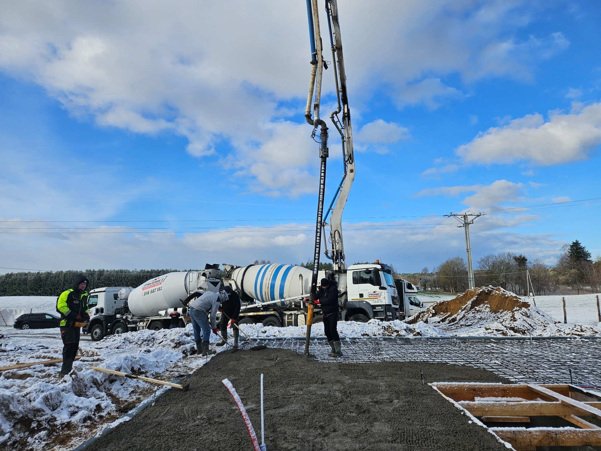 Wylewanie fundamentów domu w zimowej scenerii: ekipa budowlana rozprowadza beton za pomocą pompy, na tle ośnieżonego krajobrazu i błękitnego nieba z chmurami.