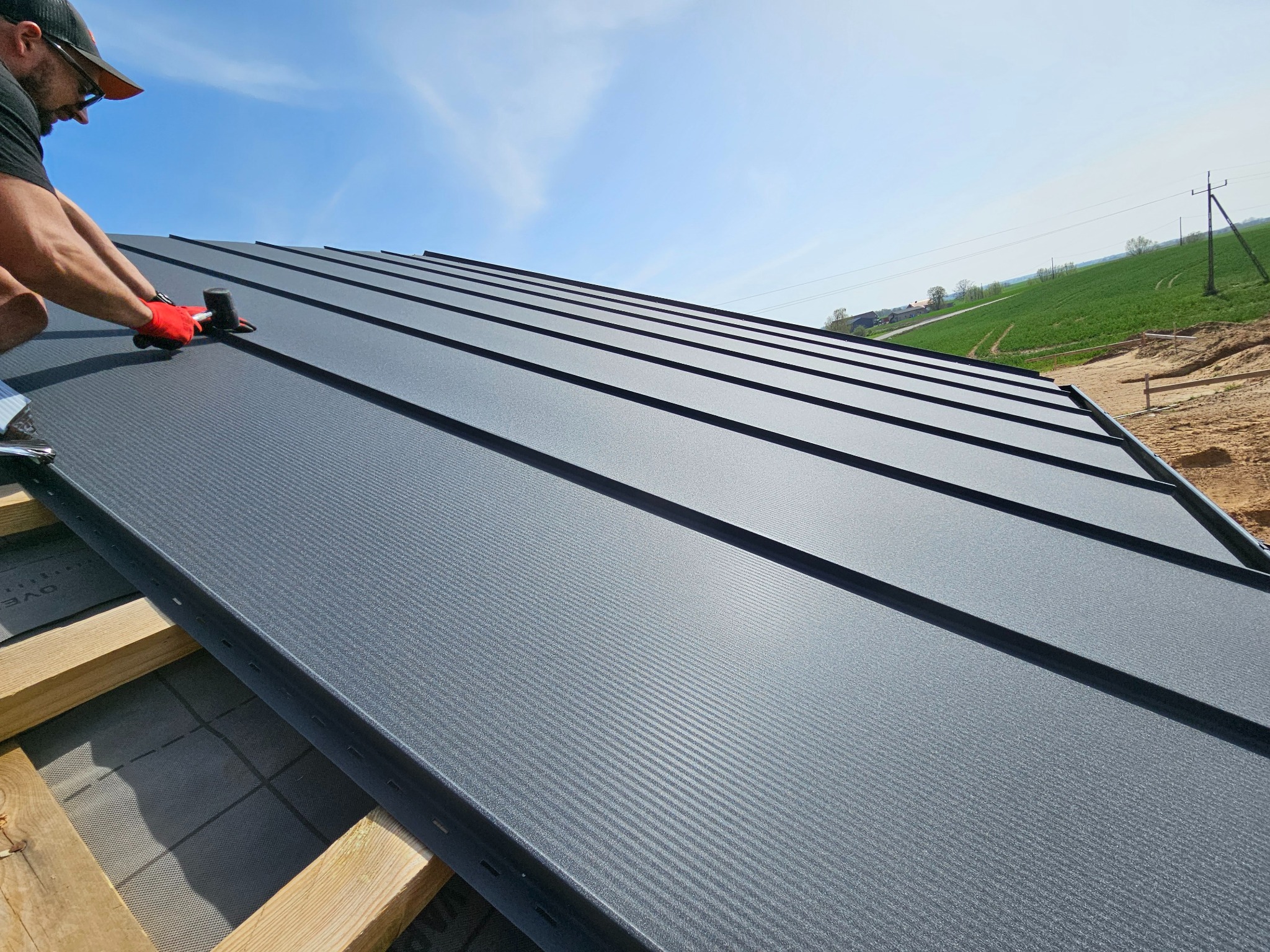 Monter w czerwonych rękawicach mocuje panel dachowy z blachy na drewnianej konstrukcji. Widok z dołu na gotowy fragment dachu i błękitne niebo w tle.