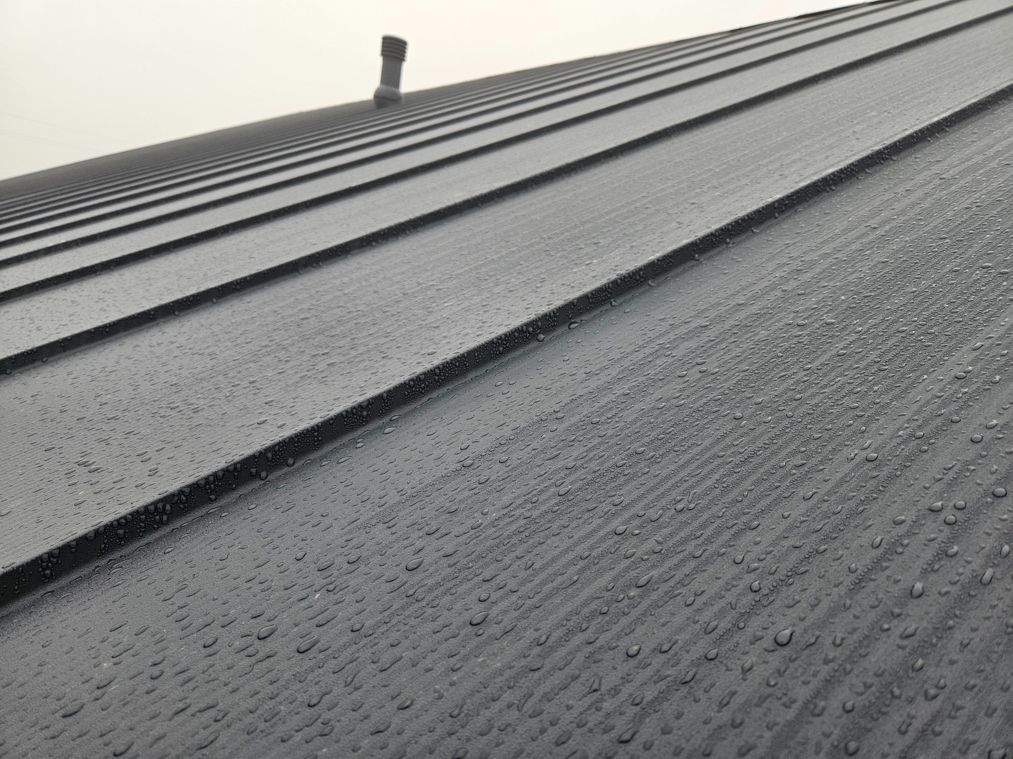 Szary, blaszany dach z widocznymi kroplami deszczu i kominkiem w tle, sfotografowany pod kątem. Widoczne regularne linie blachy dachowej.