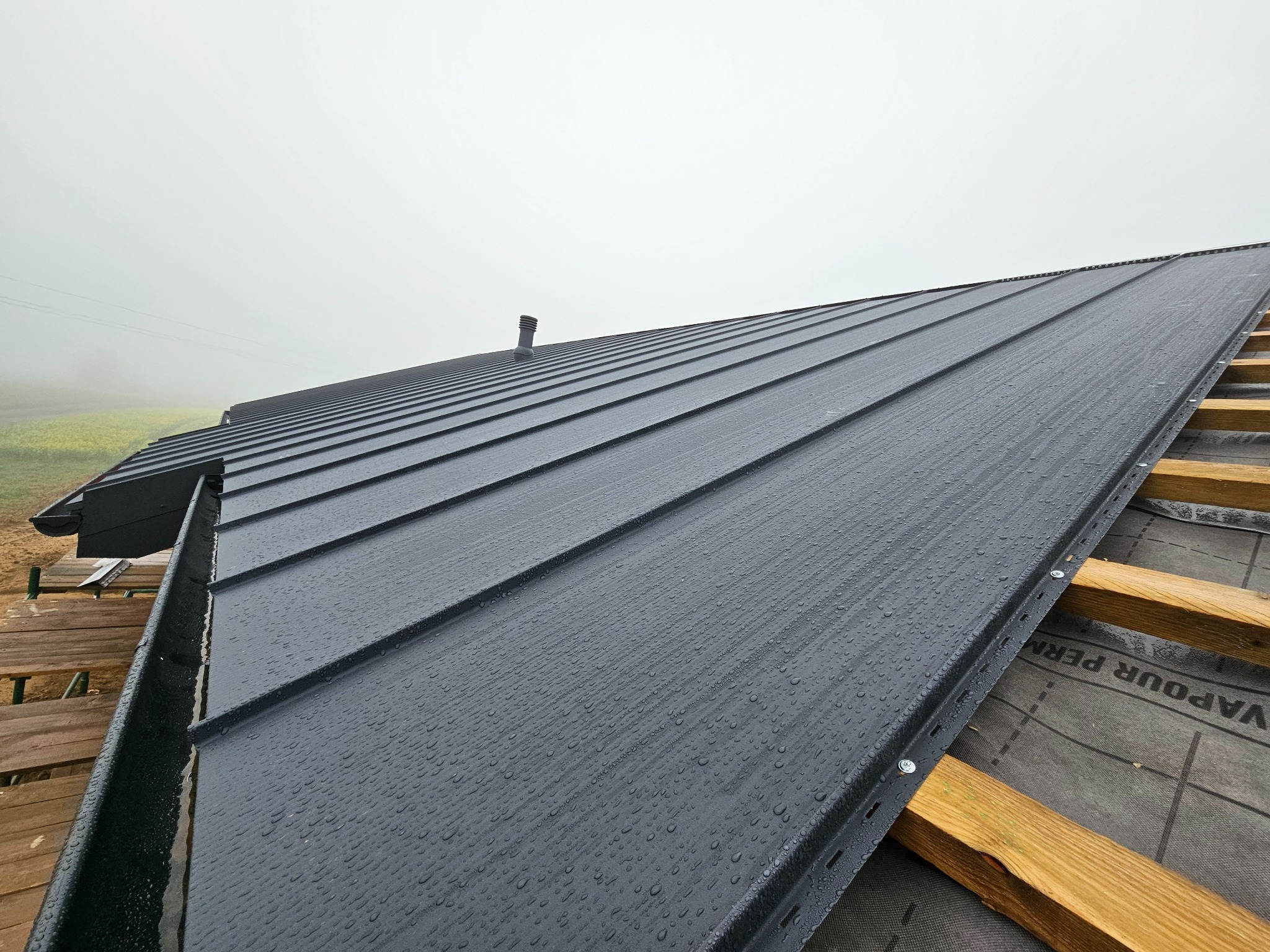 Czarny, blaszany dach z widocznymi kroplami deszczu, fragmentem rynny i drewnianą konstrukcją więźby dachowej w pochmurny dzień. Widok z dołu.