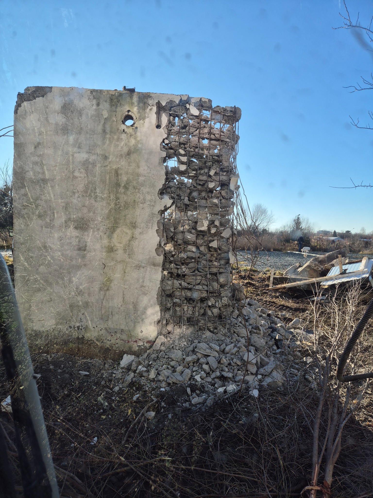 Fragment zniszczonej, betonowej ściany z odsłoniętym zbrojeniem i gruzem u podstawy, na tle błękitnego nieba i krajobrazu z drzewami. Widoczne ślady po otworze.