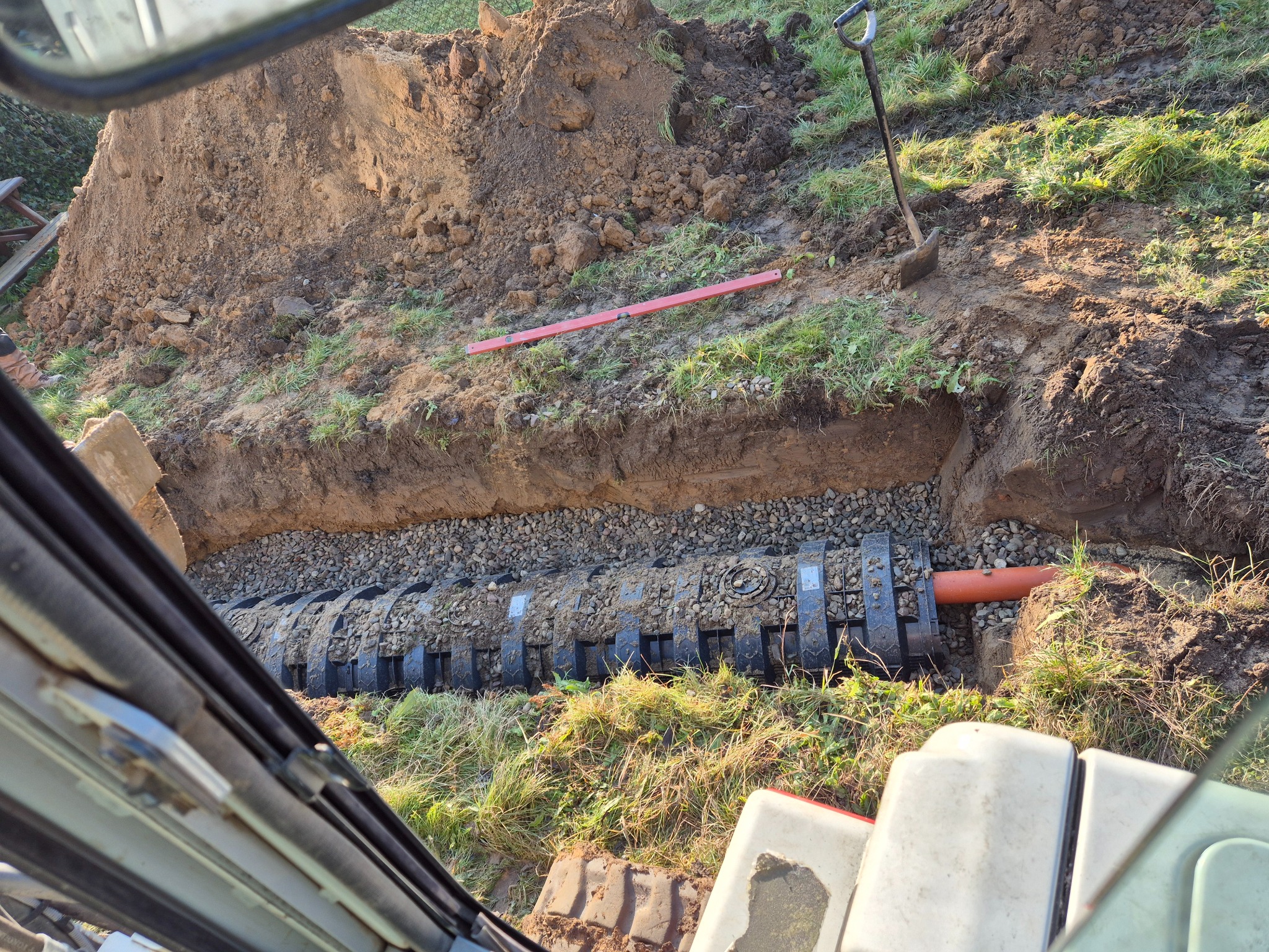Wykop pod instalacje z ułożonymi rurami drenarskimi i podsypką żwirową, widok z kabiny maszyny. Na górze wykopu leży poziomica, obok łopata.