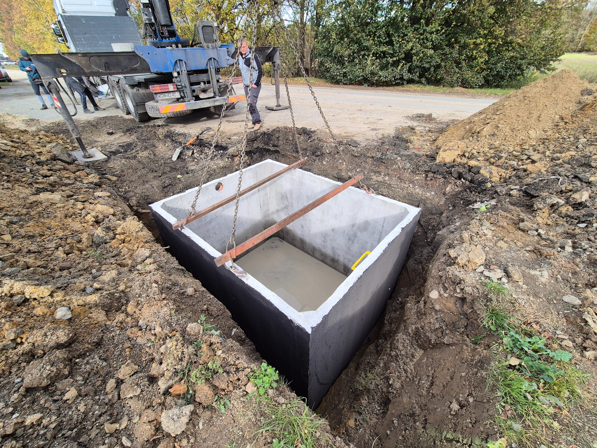 Betonowy zbiornik opuszczany do wykopu za pomocą dźwigu. Widoczny pracownik nadzorujący proces, wykopane ziemia i fragment drogi.