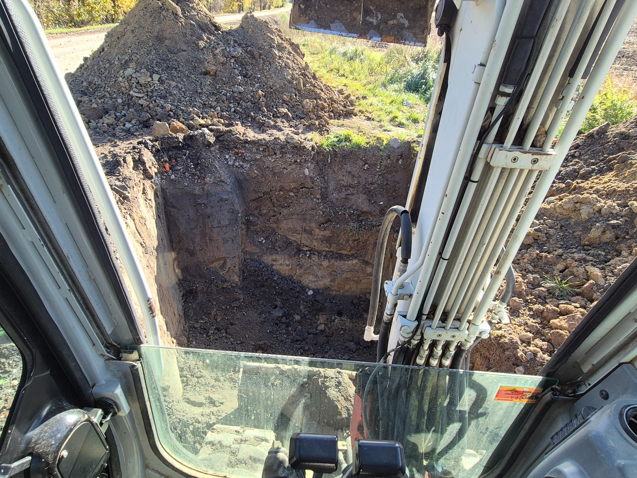 Wykopany dół z perspektywy kabiny koparki, widoczny stos ziemi i ramię maszyny. Ujęcie z wnętrza kabiny, szyba lekko zabrudzona.