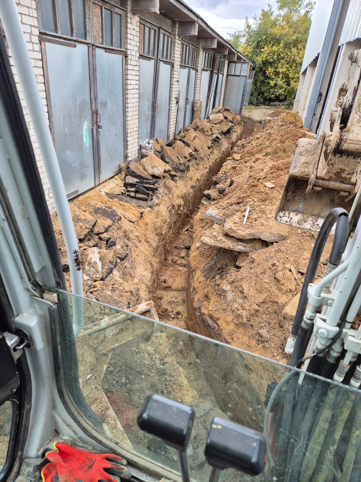 Wykopany rów w ziemi, widok z kabiny koparki. Fragment łyżki koparki widoczny po prawej. W tle budynek z szarymi bramami garażowymi i drzewa.