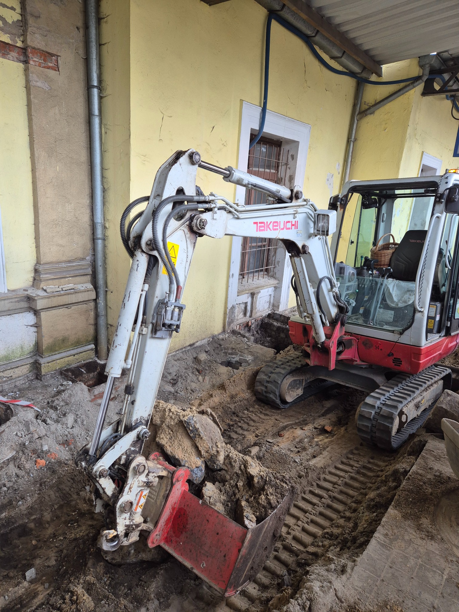 Kompaktowa koparka Takeuchi w trakcie prac ziemnych przy budynku z żółtą elewacją. Widoczne ślady gąsienic i wykopany dół. Detal: koszyk w kabinie.