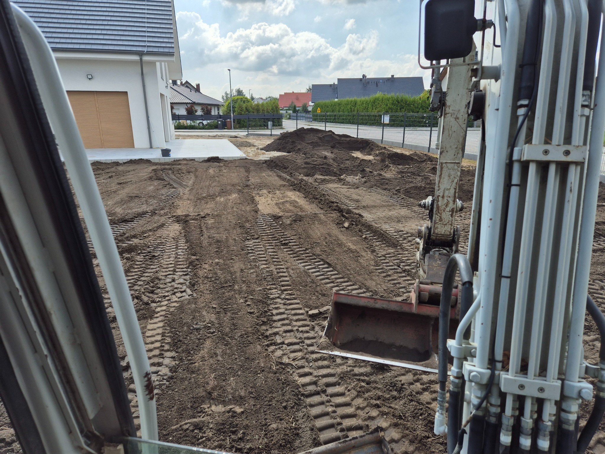 Widok z kabiny minikoparki na wyrównany teren budowy z nowym domem w tle. Ślady opon na ziemi, przygotowanie pod trawnik lub dalsze prace.