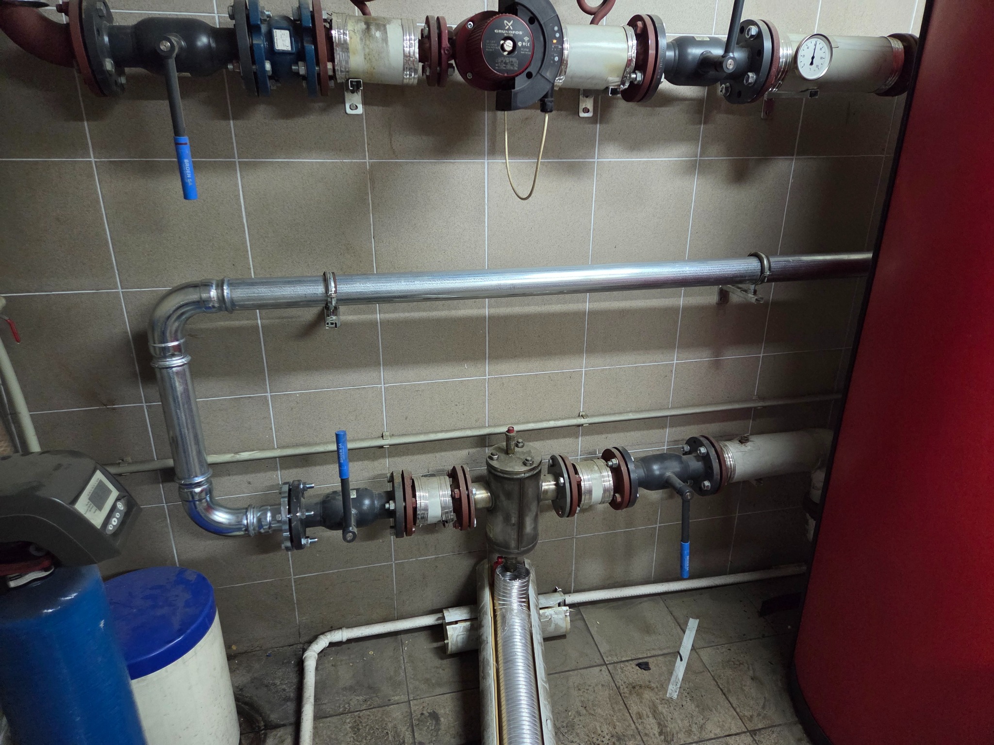 Złożona instalacja grzewcza z rurami, zaworami i pompą obiegową na tle kafelkowanej ściany. Widoczne elementy kotłowni, w tym filtr wody i czerwony zbiornik.