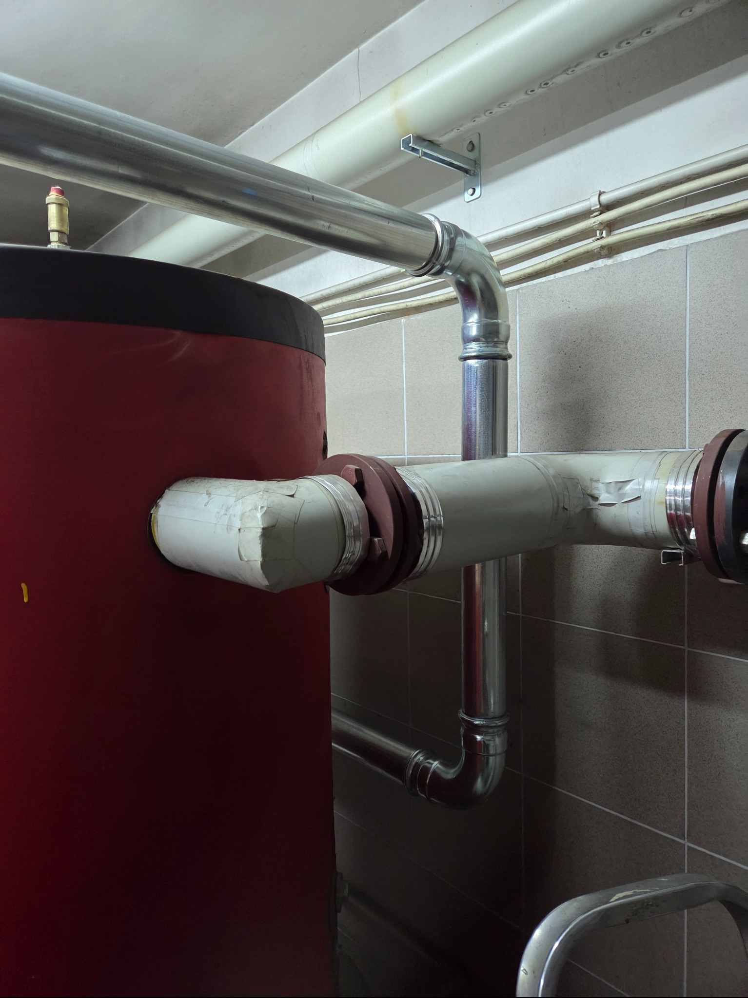 Czerwony zbiornik ciepłej wody z izolowanymi rurami i metalowymi złączkami w kotłowni. Widoczne rury biegnące wzdłuż ściany z płytek.