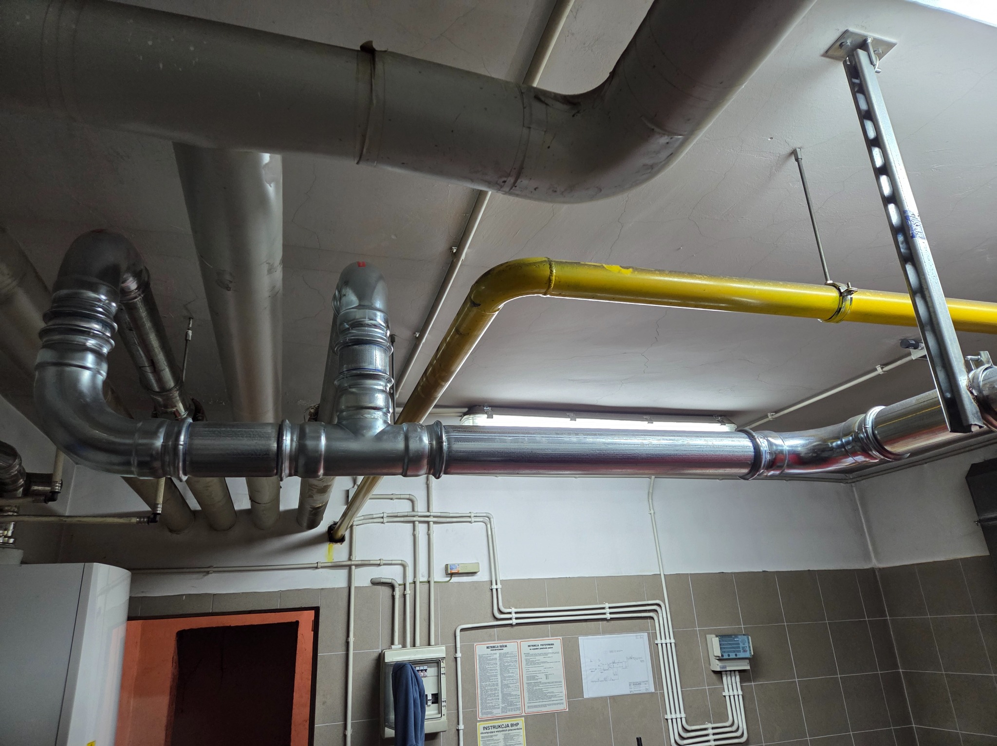 Instalacja gazowa i wodna w budynku: rury stalowe i żółte rury gazowe pod sufitem, skrzynka elektryczna, instrukcje BHP na ścianie z kafelkami.