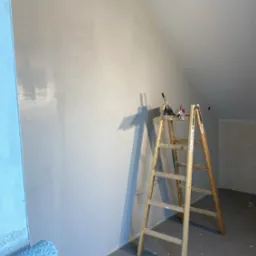 Ściana w skosie po szpachlowaniu, przygotowana do malowania. Widoczna drabina, wałek malarski i narzędzia. Powierzchnia gładka, w jasnym odcieniu szarości.