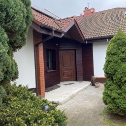 RAF-BUD - Wejście do domu z brązowymi drzwiami i elementami drewnianymi, biała elewacja, czerwona cegła, roślinność ozdobna, kostka brukowa, dachówka.