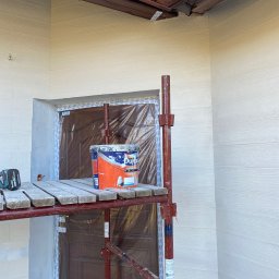 RAF-BUD - Malowanie elewacji domu z drewnopodobną fasadą. Widoczne rusztowanie, puszka farby Dulux i zabezpieczone drzwi folią malarską. W tle lampa pod okapem.