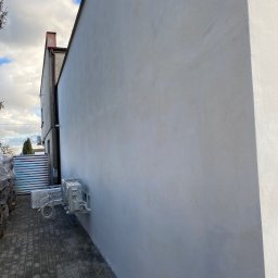 RAF-BUD - Ściana budynku po szpachlowaniu i malowaniu na szaro, z zamontowaną klimatyzacją. Widoczna kostka brukowa i materiały budowlane w tle.