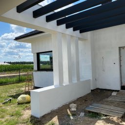 RAF-BUD - Biała elewacja domu jednorodzinnego z czarną pergolą, widok z boku, w tle zielona trawa i kukurydza. Na trawie leżą narzędzia. Częściowo widoczne rusztowanie.