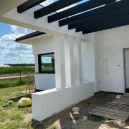 Biała elewacja domu jednorodzinnego z czarną pergolą, widok z boku, w tle zielona trawa i kukurydza. Na trawie leżą narzędzia. Częściowo widoczne rusztowanie.