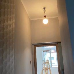 RAF-BUD - Wnętrze po odnowieniu: ściana z trójwymiarową fakturą, świeżo pomalowany sufit z lampą i drabina w przejściu do jasnego pokoju z oknem.