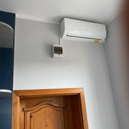 RAF-BUD - Biała klimatyzacja Hyundai na szarej ścianie nad drewnianymi drzwiami. Obok puszka elektryczna. Ujęcie z dołu, widoczny fragment szafy w kolorze granatowym.