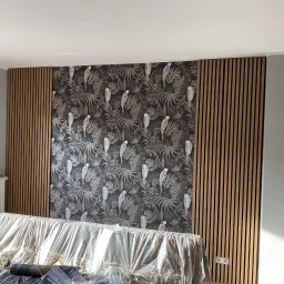 RAF-BUD - Ściana z panelami akustycznymi i tapetą w tropikalne wzory, częściowo zakryta folią ochronną. Widoczny grzejnik i drabina malarska.