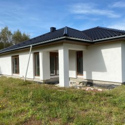 RAF-BUD - Biały dom parterowy z ciemną dachówką w słoneczny dzień. Widoczne okna z brązowymi ramami, kolumny werandy i fragment dachu. Tło stanowią drzewa i błękitne niebo.