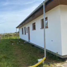 Świeżo otynkowany, parterowy dom z brązowymi oknami i szarymi rynnami, otoczony zieloną trawą i częściowo widocznym ogrodzeniem. Widoczne detale wykończenia elewacji.