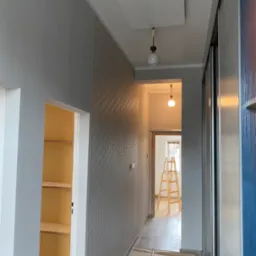 Korytarz po remoncie: szara ściana z fakturą, białe drzwi do garderoby z półkami, w tle drabina malarska. Widoczne detale oświetlenia i wykończenia.