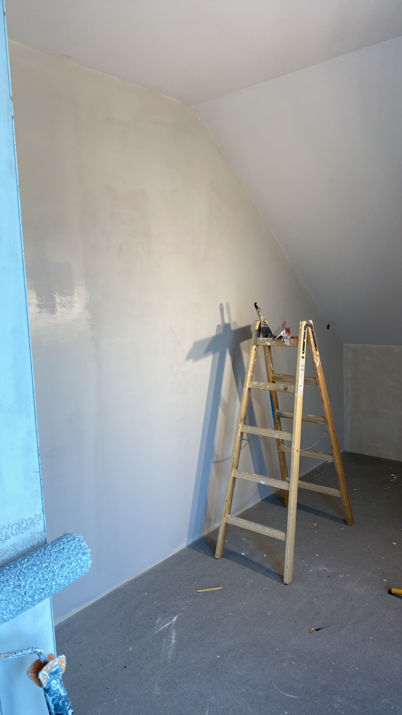 Ściana w skosie po szpachlowaniu, przygotowana do malowania. Widoczna drabina, wałek malarski i narzędzia. Powierzchnia gładka, w jasnym odcieniu szarości.