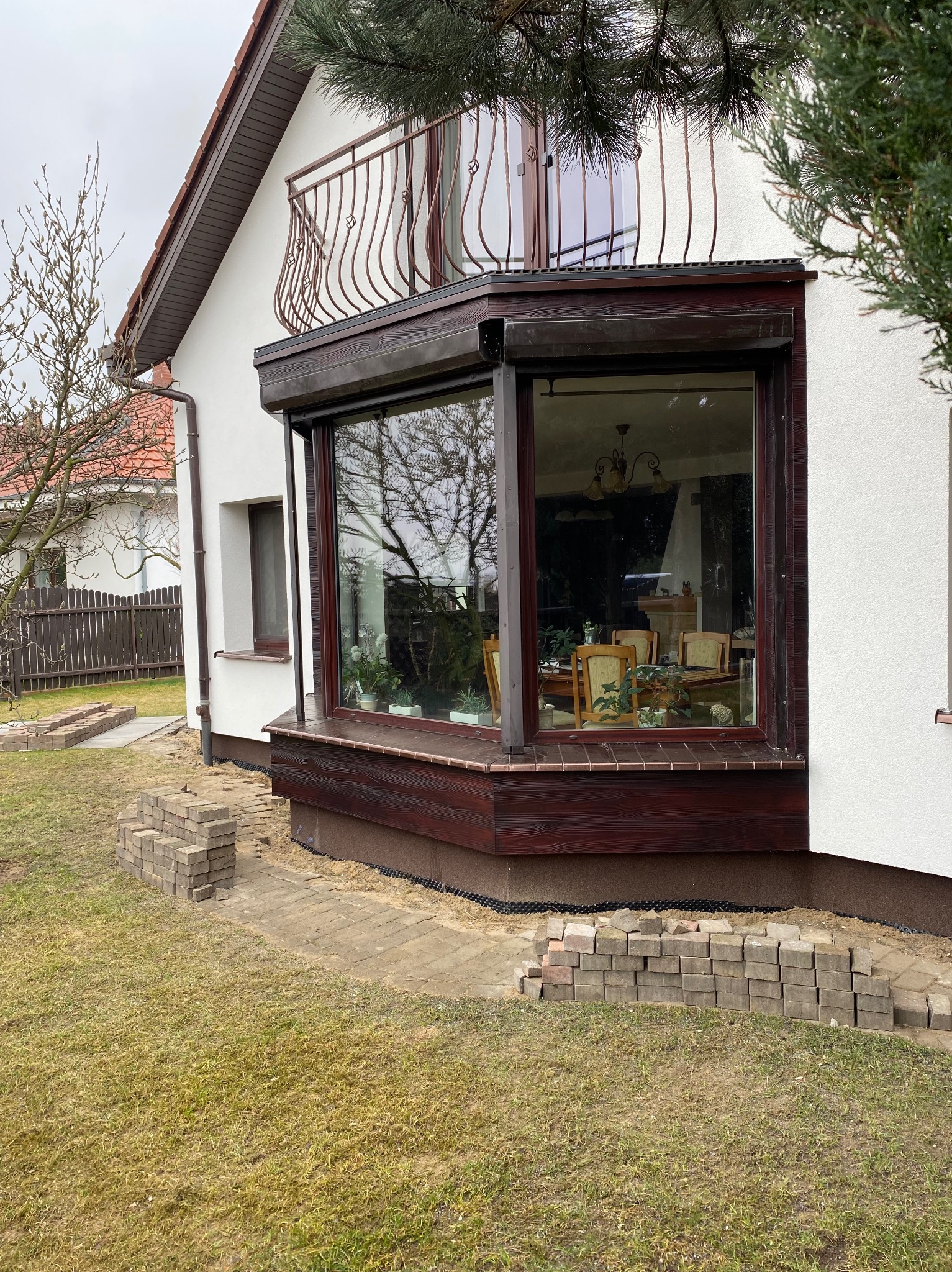 Biały dom z brązowymi akcentami: okna z roletami, balkon z kutą balustradą. Ułożona kostka brukowa wokół budynku. Widok na wnętrze domu przez duże okno.