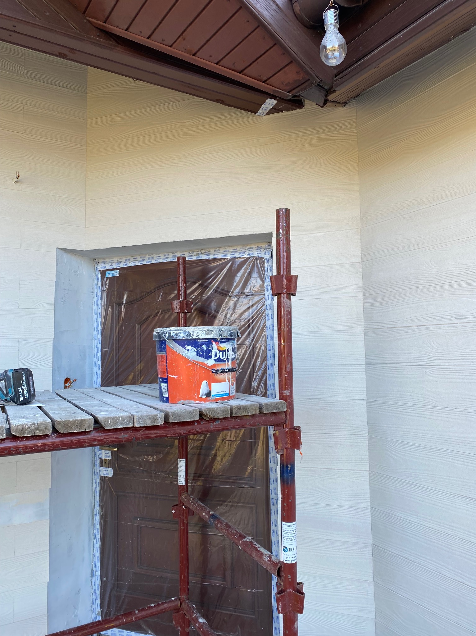 Malowanie elewacji domu z drewnopodobną fasadą. Widoczne rusztowanie, puszka farby Dulux i zabezpieczone drzwi folią malarską. W tle lampa pod okapem.