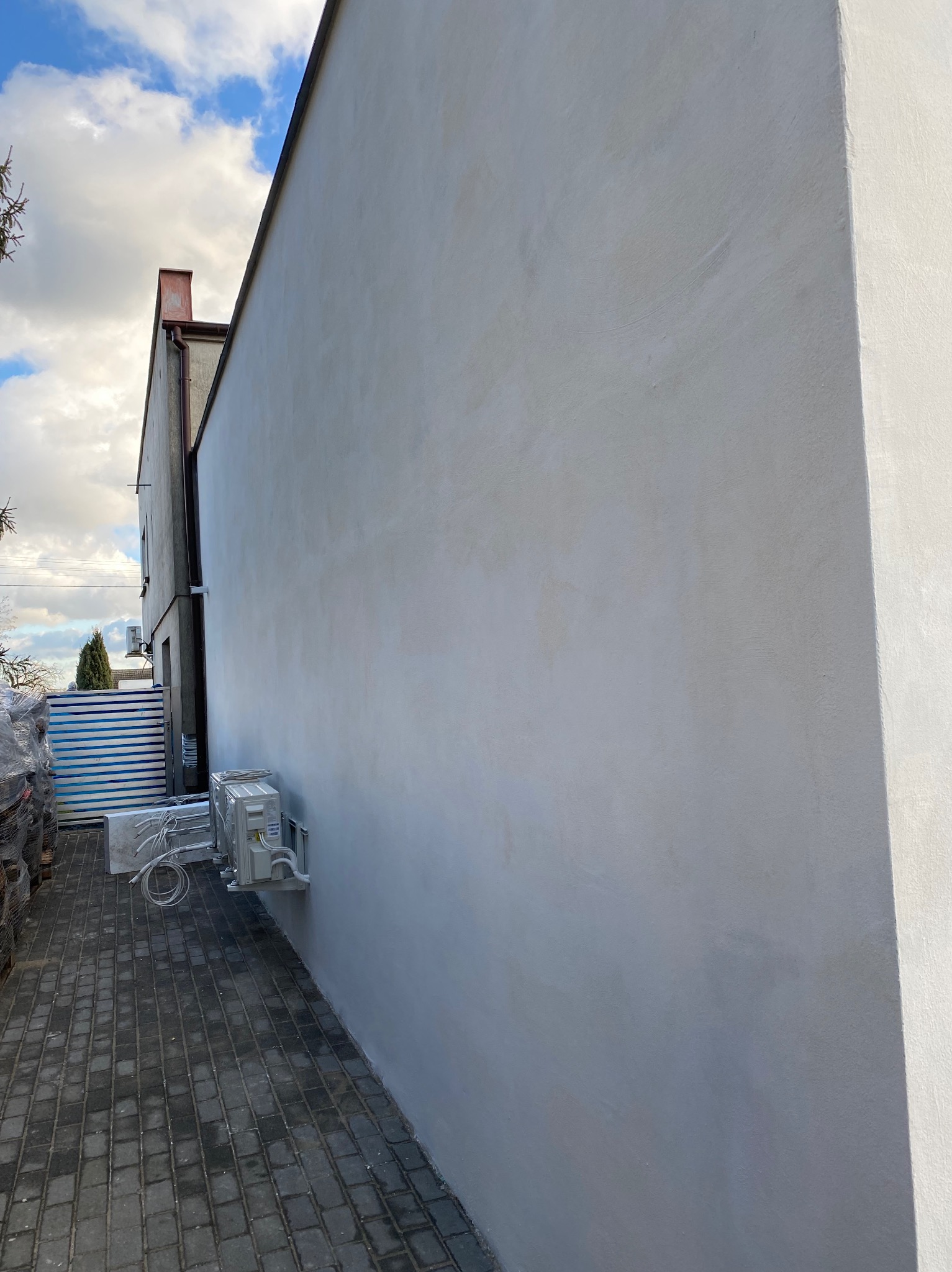 Ściana budynku po szpachlowaniu i malowaniu na szaro, z zamontowaną klimatyzacją. Widoczna kostka brukowa i materiały budowlane w tle.