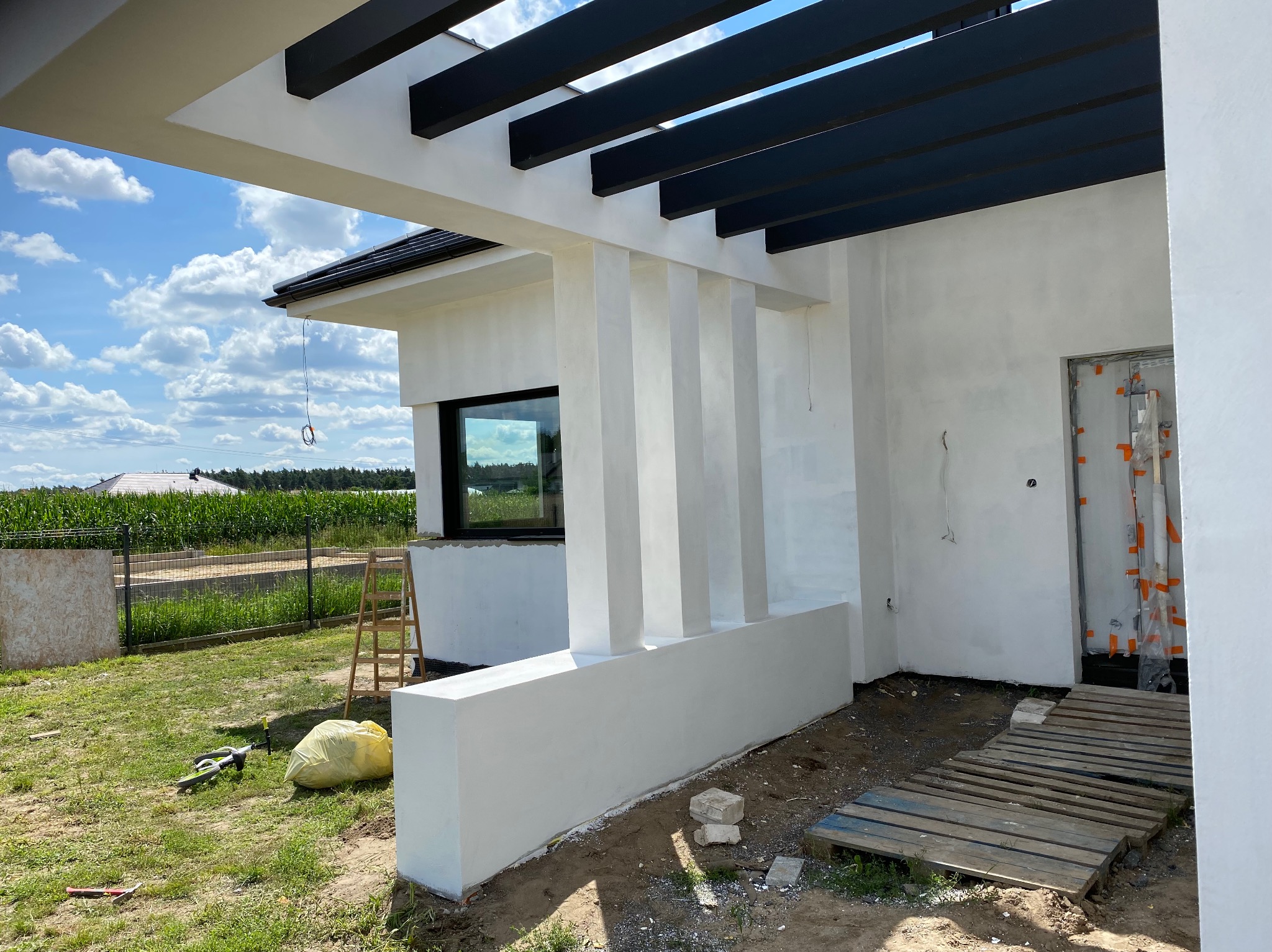 Biała elewacja domu jednorodzinnego z czarną pergolą, widok z boku, w tle zielona trawa i kukurydza. Na trawie leżą narzędzia. Częściowo widoczne rusztowanie.