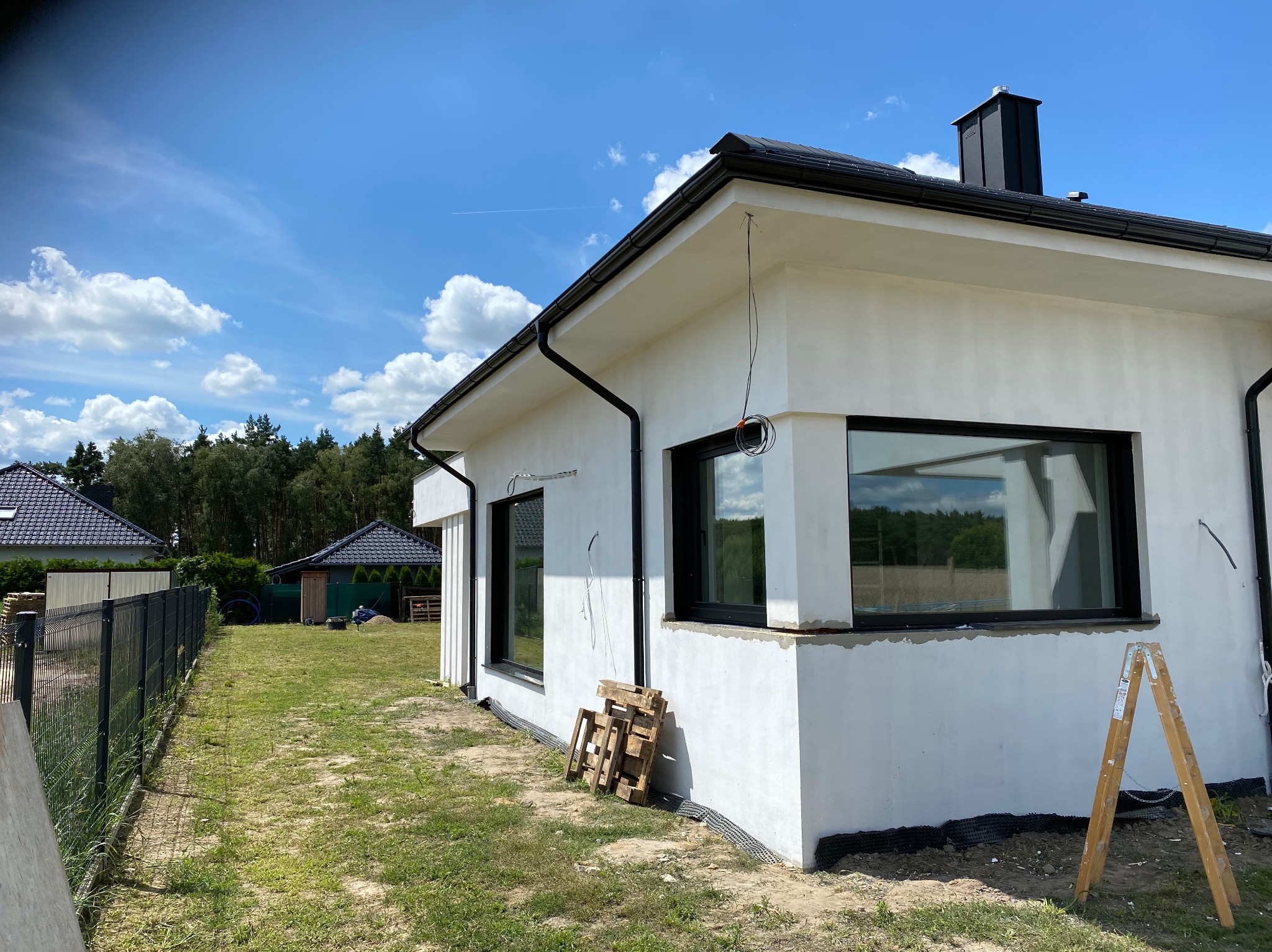 Boczny widok domu w trakcie prac elewacyjnych, z widocznymi oknami w czarnych ramach, fragmentem rusztowania i przygotowanym podłożem. Jasne tło nieba.