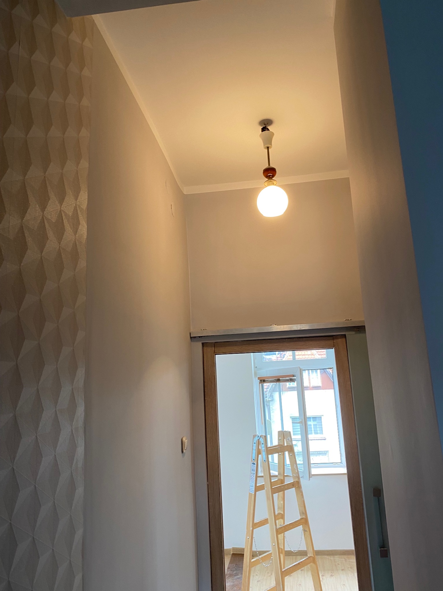 Wnętrze po odnowieniu: ściana z trójwymiarową fakturą, świeżo pomalowany sufit z lampą i drabina w przejściu do jasnego pokoju z oknem.