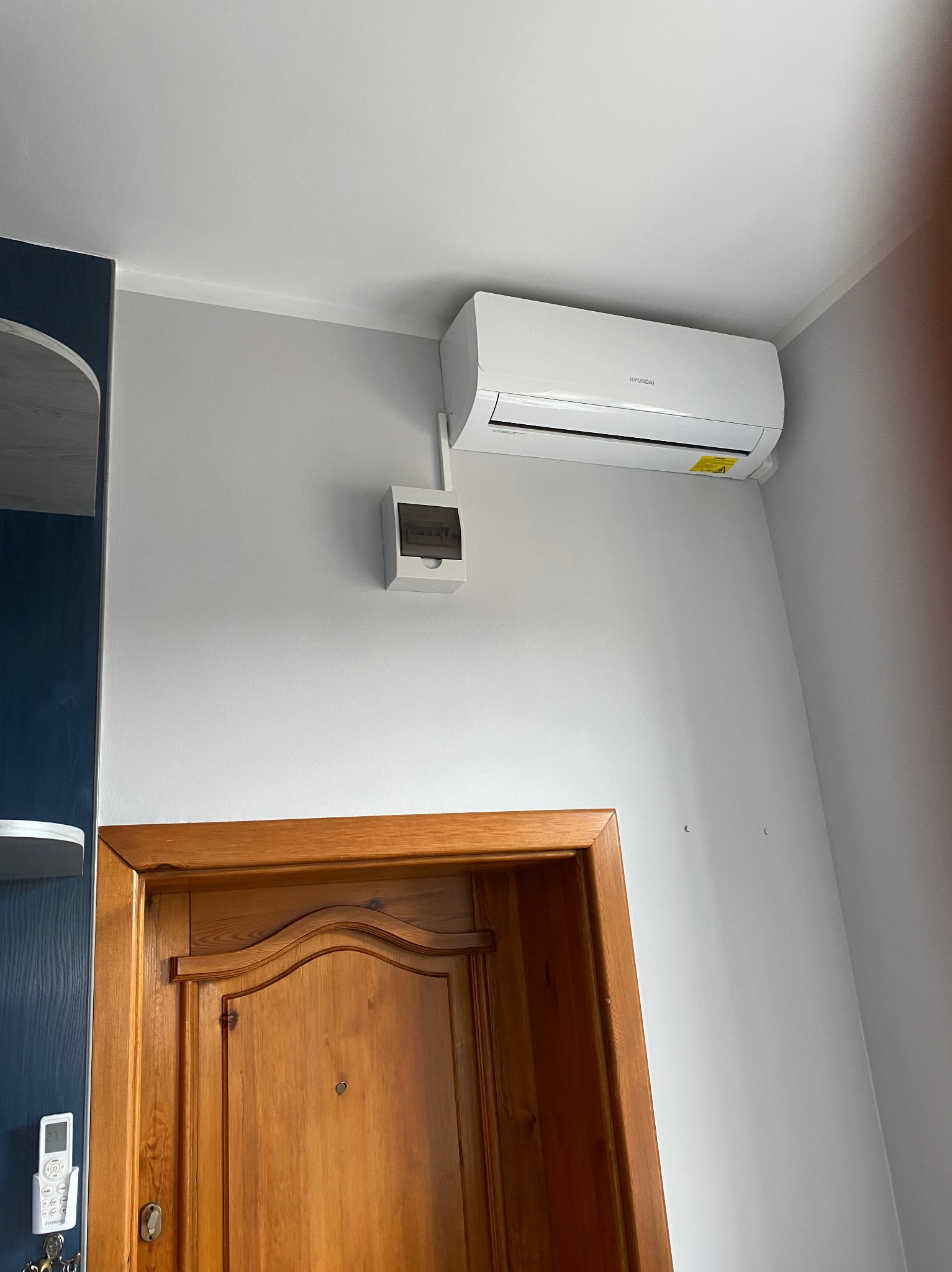 Biała klimatyzacja Hyundai na szarej ścianie nad drewnianymi drzwiami. Obok puszka elektryczna. Ujęcie z dołu, widoczny fragment szafy w kolorze granatowym.