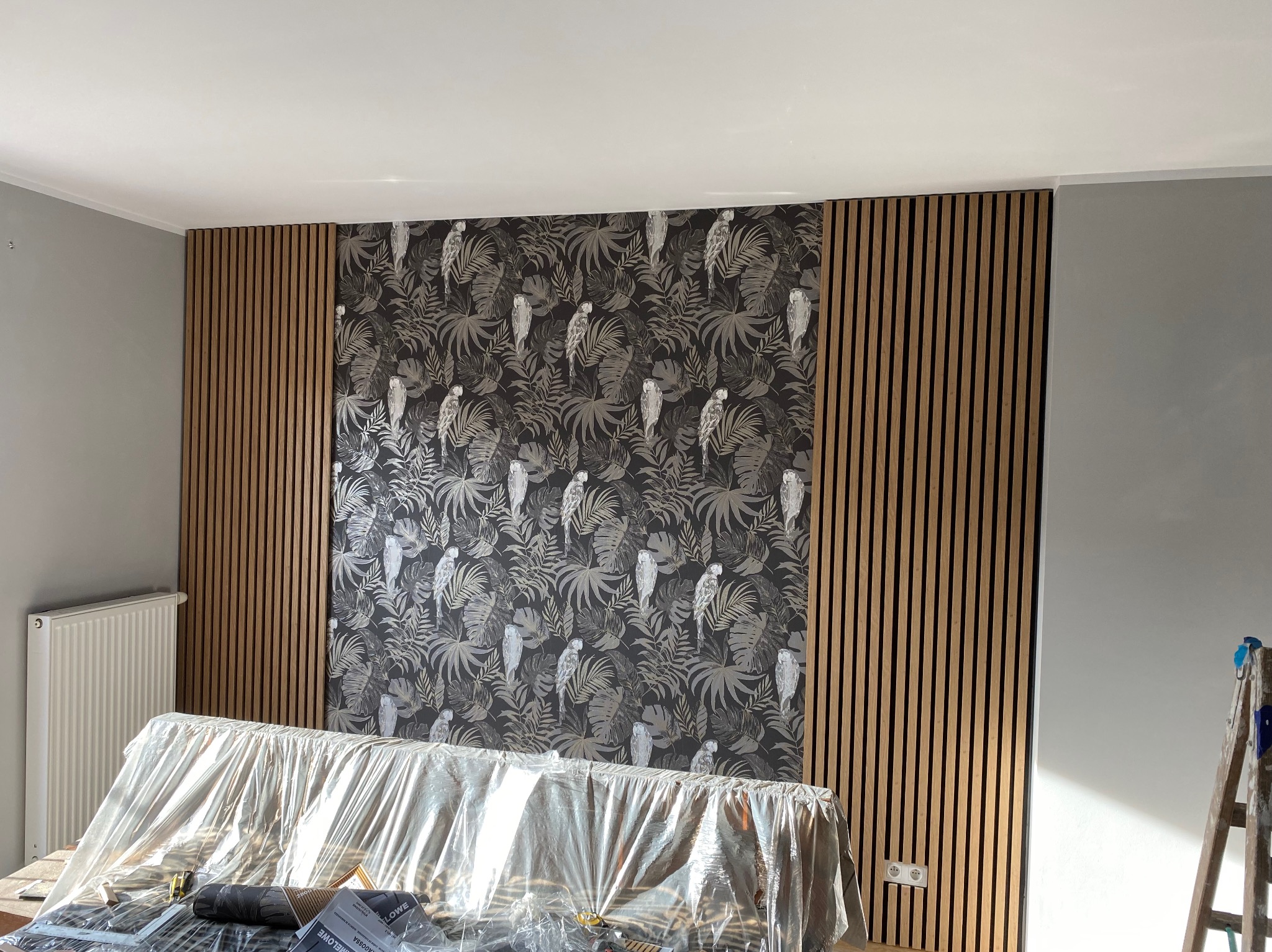 Ściana z panelami akustycznymi i tapetą w tropikalne wzory, częściowo zakryta folią ochronną. Widoczny grzejnik i drabina malarska.