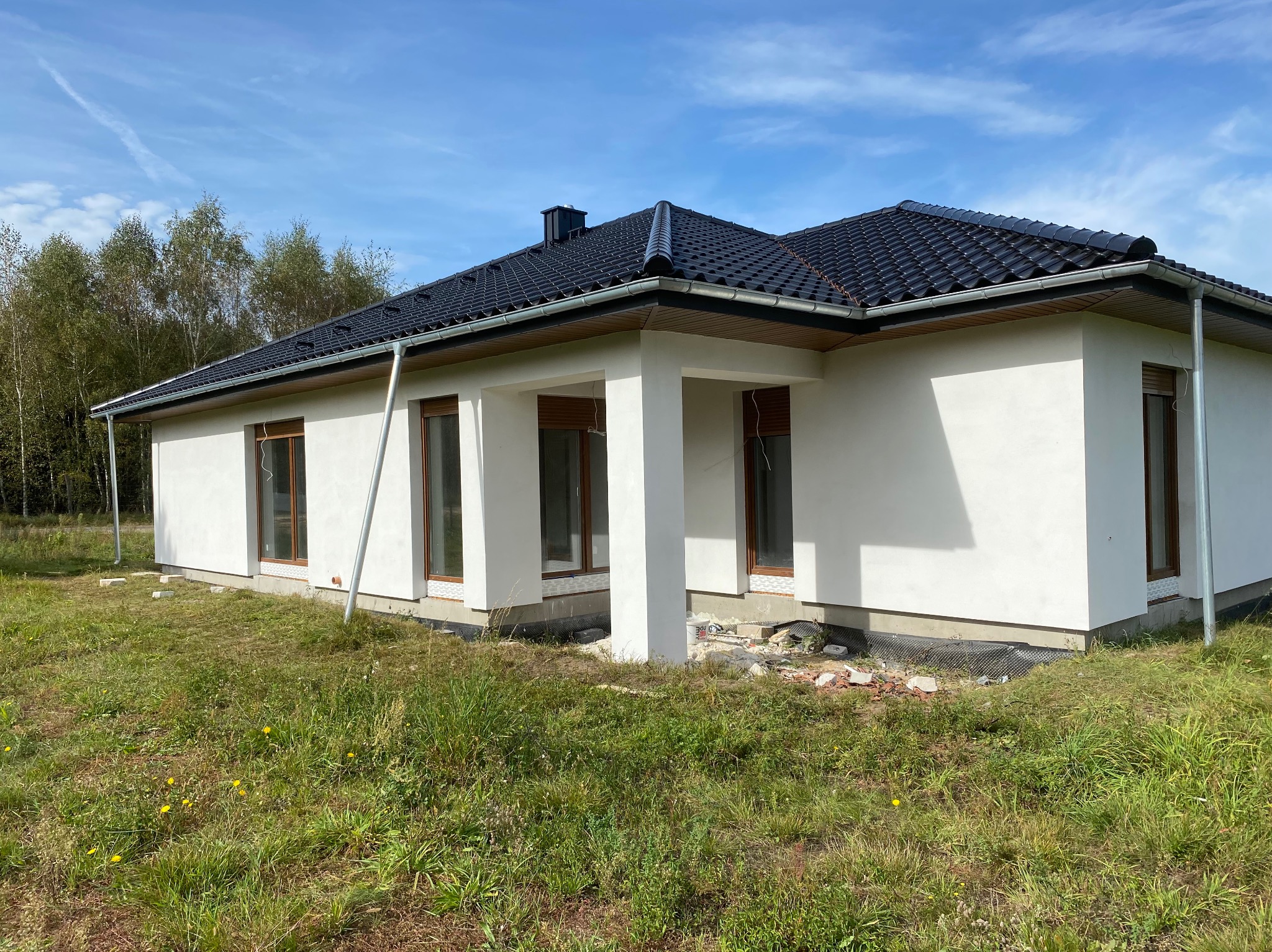 Biały dom parterowy z ciemną dachówką w słoneczny dzień. Widoczne okna z brązowymi ramami, kolumny werandy i fragment dachu. Tło stanowią drzewa i błękitne niebo.