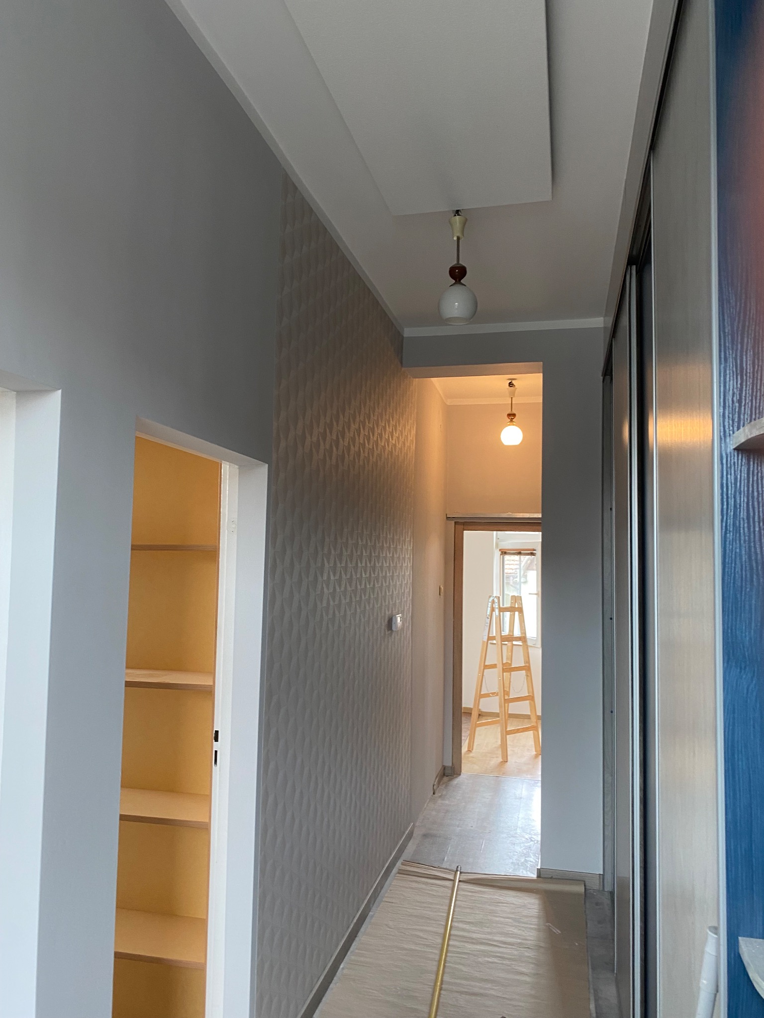 Korytarz po remoncie: szara ściana z fakturą, białe drzwi do garderoby z półkami, w tle drabina malarska. Widoczne detale oświetlenia i wykończenia.
