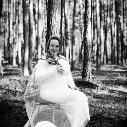 Weronika Wojtczak - Czarno-białe zdjęcie kobiety w białej sukience i welonie siedzącej na krześle w lesie, trzymającej mały bukiet kwiatów.