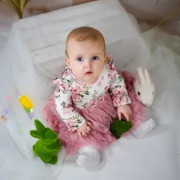 Niemowlę w kwiecistej sukience i tiulowej spódniczce siedzi na białym tle z dekoracjami wielkanocnymi, w tym pluszowym króliczkiem i zielonymi zajączkami z mchu.