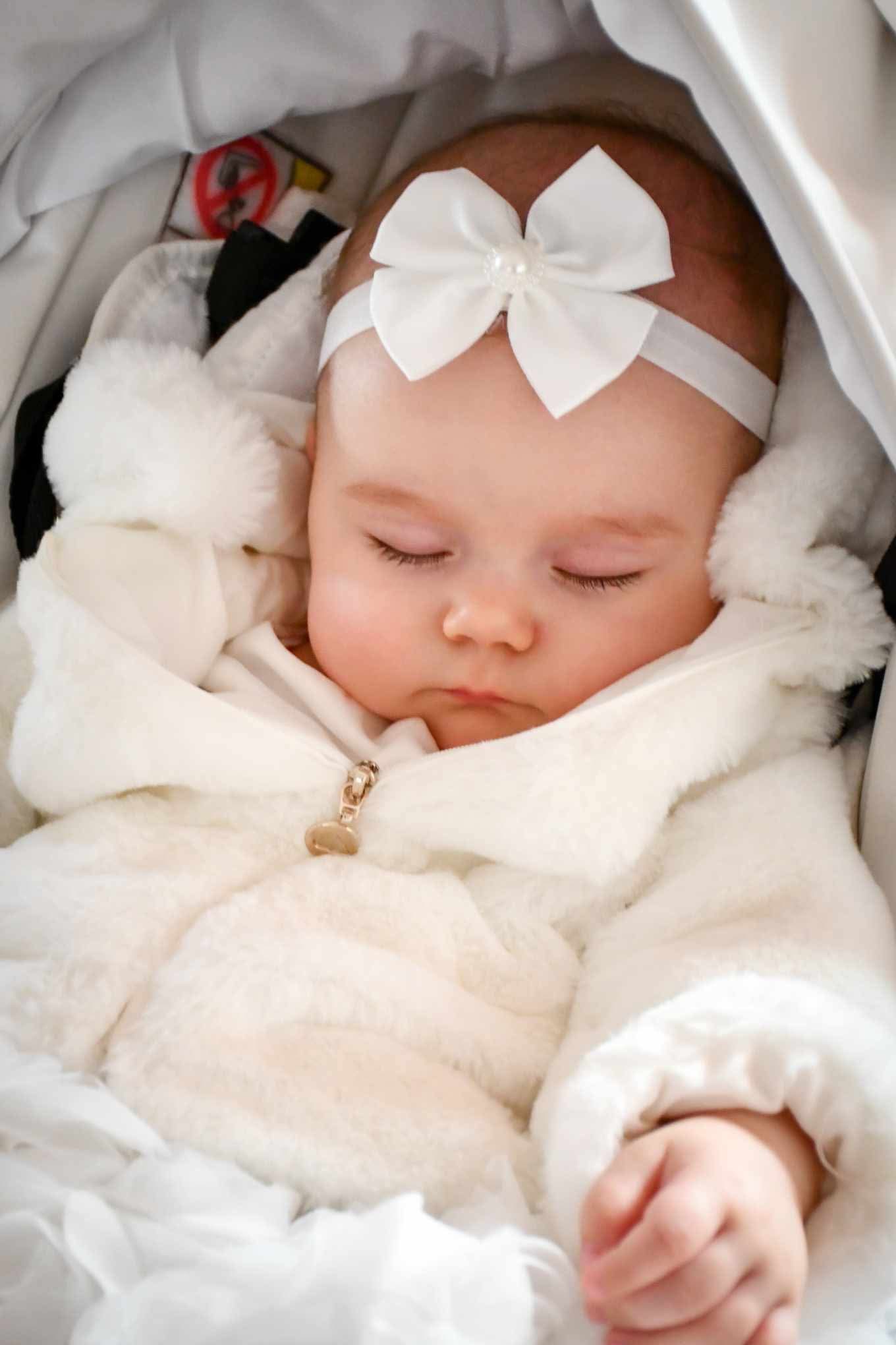 Śpiąca niemowlę w białym futrzanym kombinezonie z kokardą na głowie, widoczny zamek błyskawiczny w kształcie serca i mała rączka.