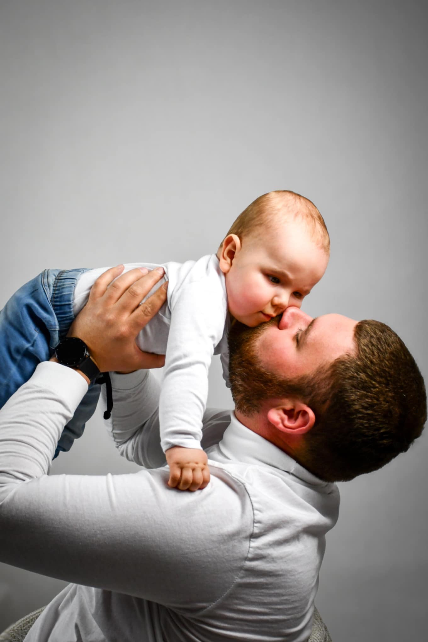 Ojciec z brodą trzymający niemowlę w dżinsach, zbliżający się nosem do jego noska na tle szarego tła.