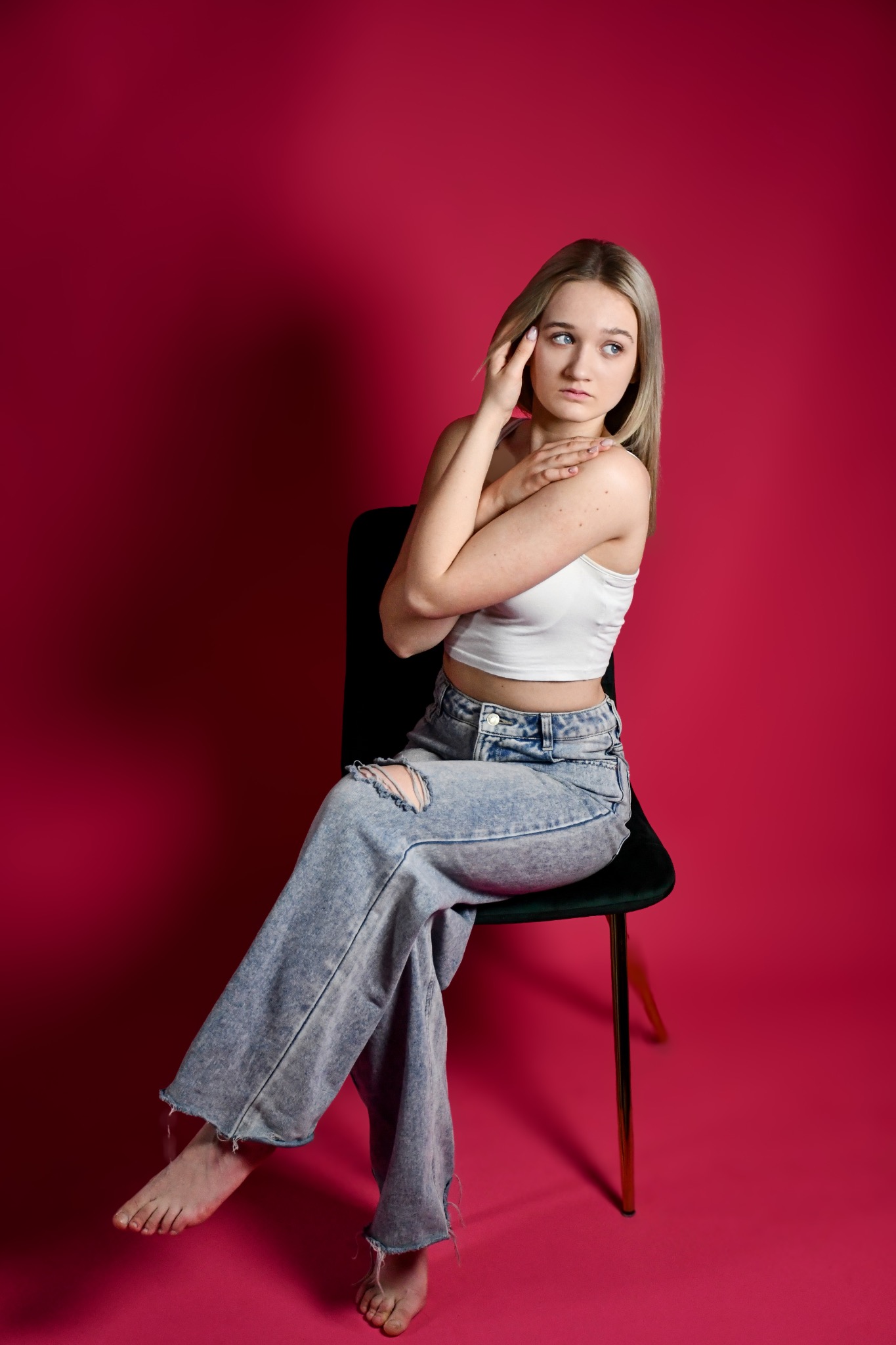 Młoda kobieta z blond włosami siedzi na krześle na tle fuksjowego studia, ubrana w biały top i podarte dżinsy, zamyślona spoglądając w lewo.