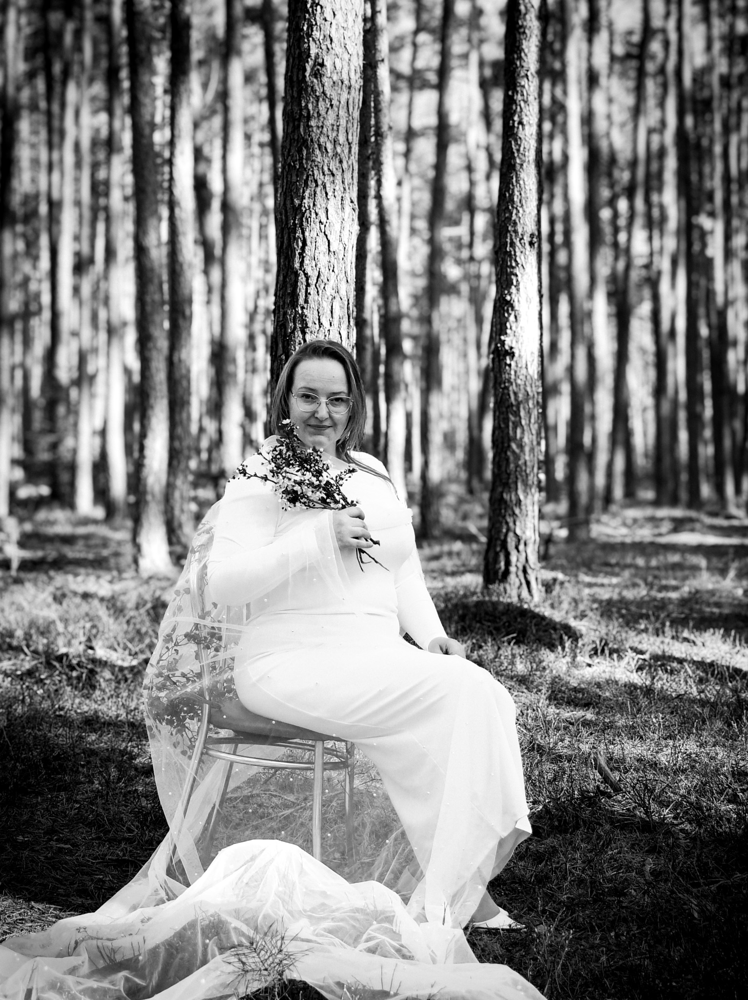 Czarno-białe zdjęcie kobiety w białej sukience i welonie siedzącej na krześle w lesie, trzymającej mały bukiet kwiatów.