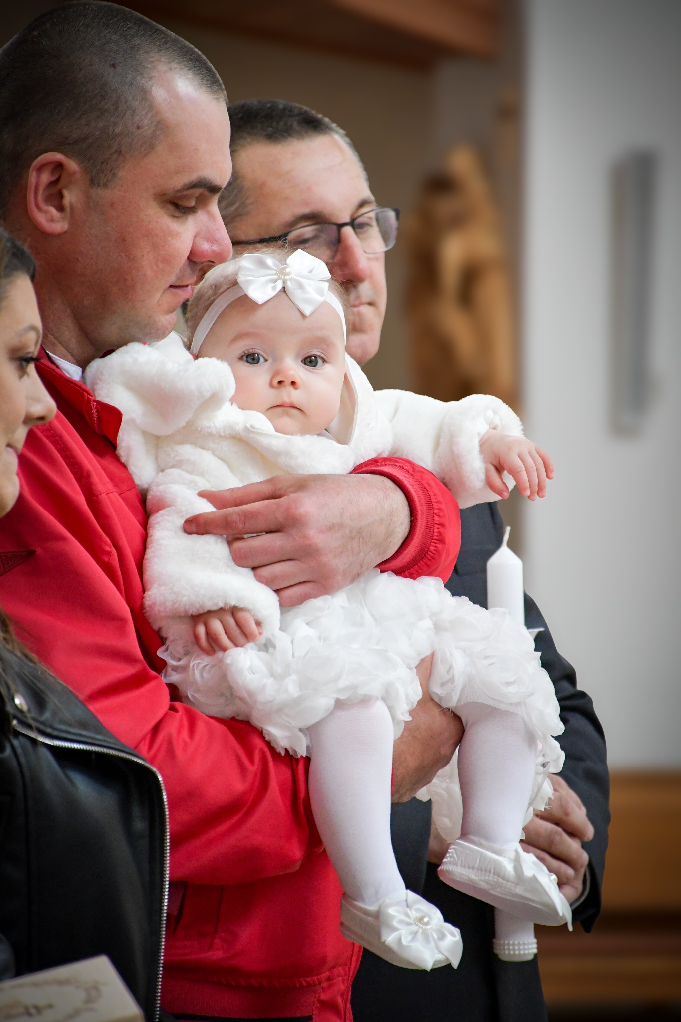 Uroczyste chrzciny: Dziewczynka w białym stroju z kokardą na głowie, trzymana przez mężczyznę w czerwonej kurtce, obok stoi mężczyzna w okularach, w tle widać świecę i elementy drewniane.